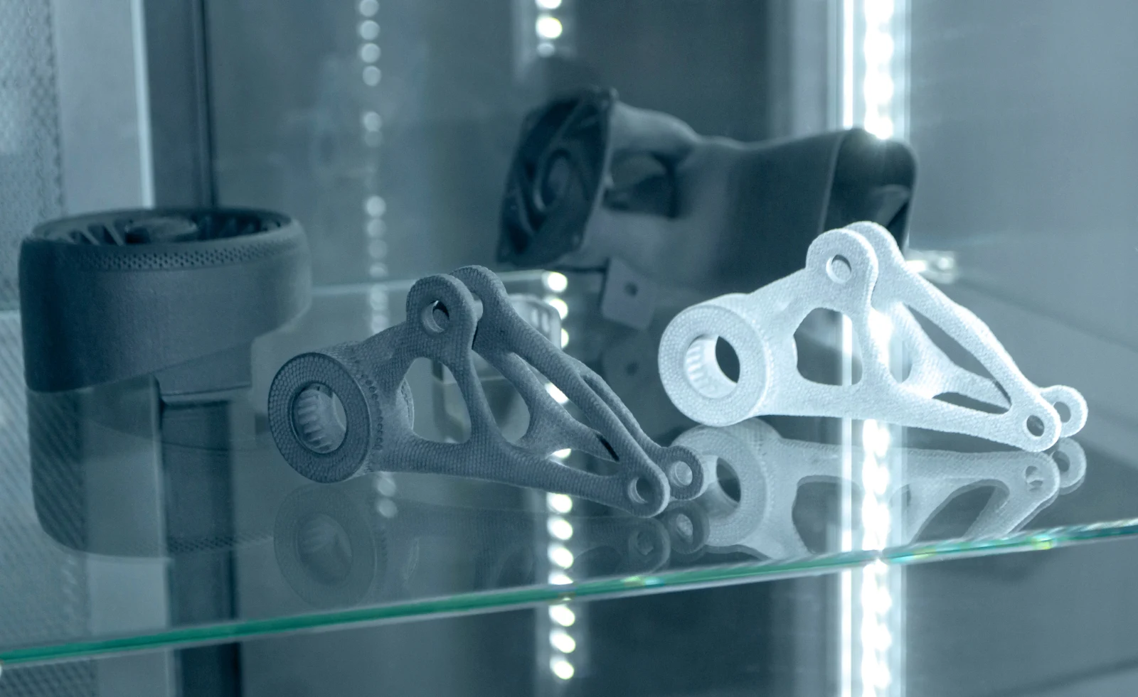 Two topology-optimised metal brackets displayed on a lit glass shelf