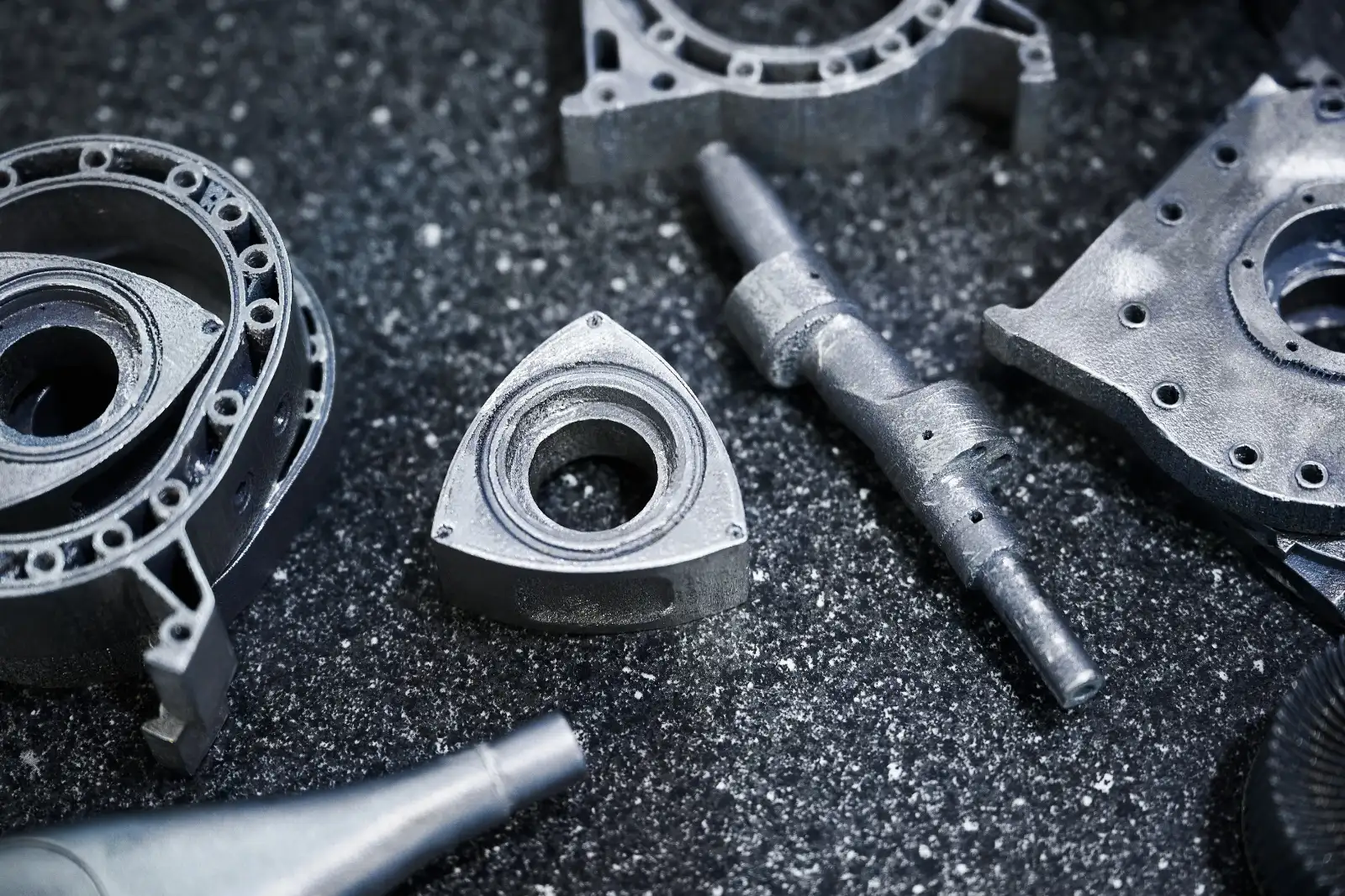 Machined and printed metal engine components laid out on a dark speckled surface